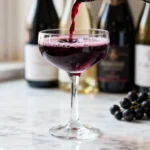Verre de kir en gros plan avec vin blanc aligoté et liqueur de cassis versés, bouteilles et fruits en arrière-plan flou