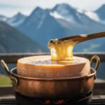 Raclette du Valais AOP fondant dans un pot en cuivre traditionnel, avec croûte dorée et grattoir en bois devant des montagnes suisses