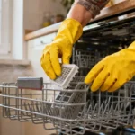 Femme portant des gants jaunes nettoyant le filtre d'un lave-vaisselle dans une cuisine éclairée naturellement
